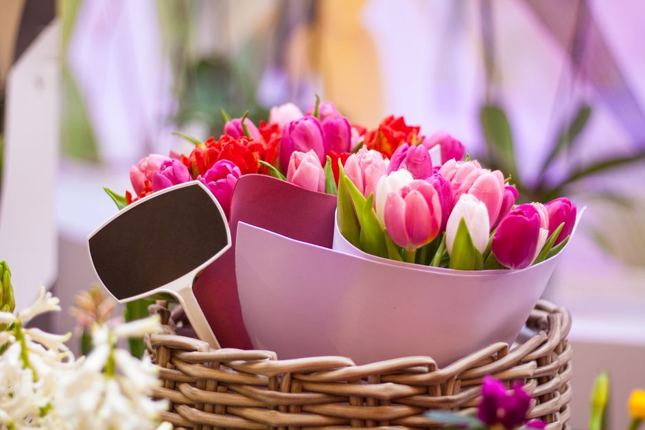 A vibrant floral arrangement featuring a mix of pink and white tulips with green stems and leaves, arranged in a sleek, white, curved ceramic bowl. The bouquet is complemented by red and light pink fl