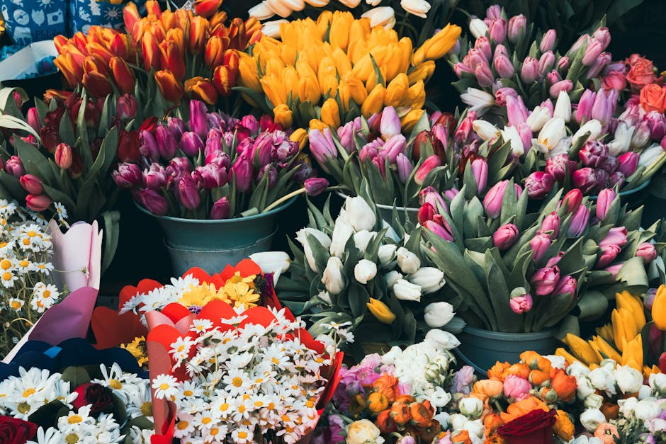 A vibrant collection of fresh flowers arranged in various bouquets and containers. Prominent in the image are numerous tulips in shades of pink, white, yellow, and orange, with some still in bud form