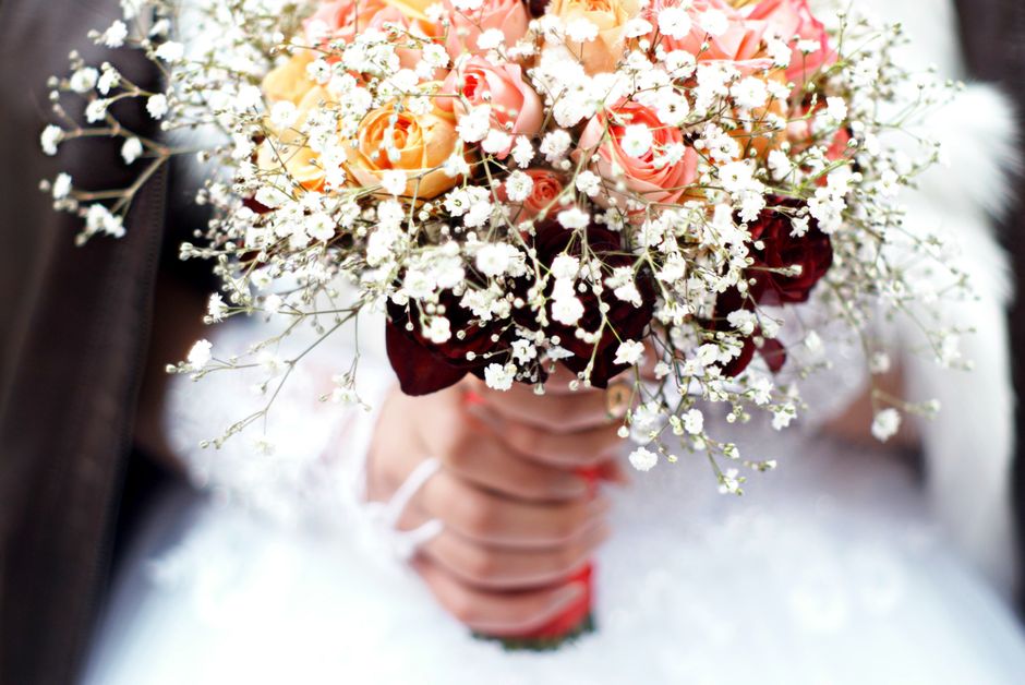 A close-up of a bridal bouquet featuring soft pink and peach roses, interspersed with delicate white baby's breath flowers. The bouquet is wrapped in dark burgundy paper with a ribbon, held by a bride