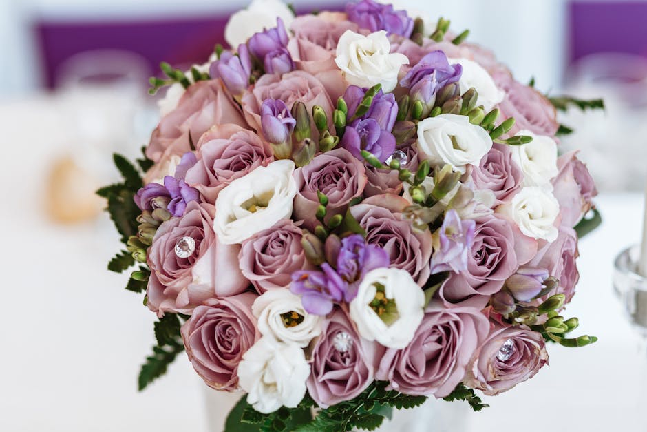 A close-up of a bouquet arrangement consisting of light pink roses, white lisianthus, and purple freesia, accented with small green foliage and decorative rhinestones nestled among the flowers. The bo