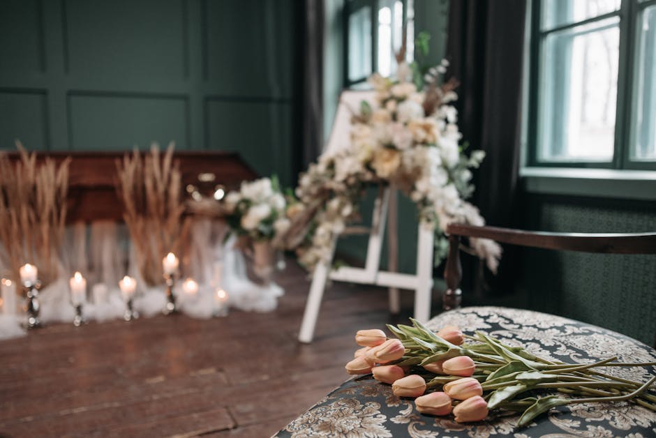 Close-up view of a bouquet of soft pink tulips with green stems resting on a patterned fabric surface, set in a room with wooden flooring and dark green paneled walls. In the background, there is an a
