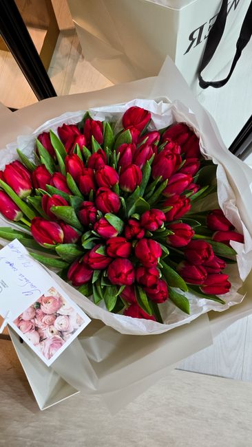 A large bouquet of fresh red tulips with green stems and leaves, arranged in a dense cluster and wrapped in white tissue paper. The tulips have smooth, slightly curved petals that are vibrant and glos