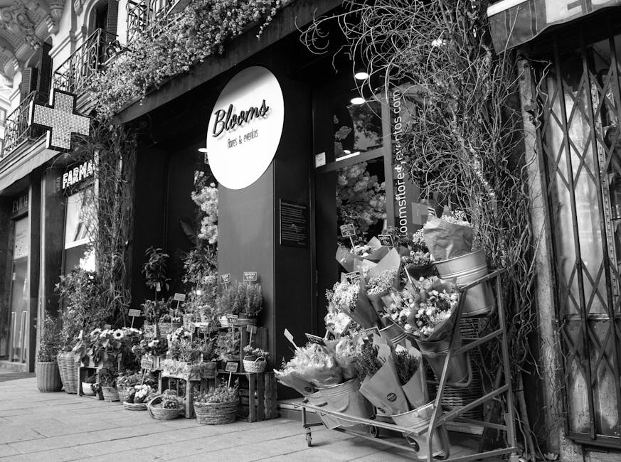 A street view of a flower shop named 'Bloom' with a sign reading 'flowers & gifts' on a modern storefront. The entrance is adorned with an array of fresh floral arrangements and potted plants placed o