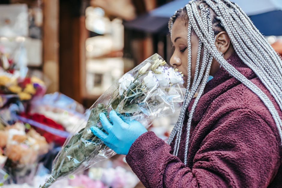 A woman with silver braided hair is seen at a flower market, holding a bouquet of white roses wrapped in clear plastic. She is wearing a maroon fleece jacket and bright blue gloves, closely inspecting
